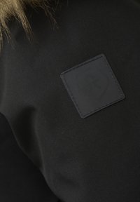 Svart ytterplagg med en texturerad tyg och en framträdande svart läderpatch med en stiliserad logotyp. Fuskpäls vid kragen.