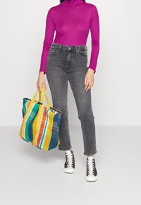 Kolorowa torba na ramię w paski z żółtymi uchwytami, zestawiona z dopasowaną fioletową bluzką z długim rękawem i szarymi dżinsami z wysokim stanem. Czarne trampki.