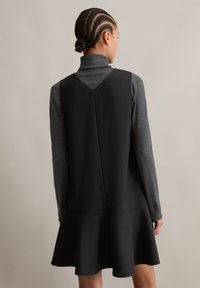 Μαύρο φόρεμα χωρίς μανίκια με εφαρμοστή κορσέ και φουσκωτή φούστα, που διαθέτει υφασμάτινη υφή και βαθύ V-ανοιχτό πίσω μέρος, επενδυμένο με γκρι πουλόβερ με ψηλό λαιμό.