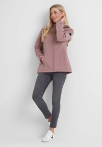 Soft shell jacket in dusty pink, with a high neck, zip closure, and two front pockets. Paired with grey skinny jeans and white sneakers.