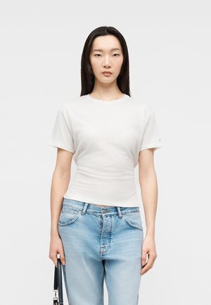 Femme aux longs cheveux foncés portant un t-shirt blanc uni à manches courtes et un jean bleu clair, tenant un sac à main noir à côté d'elle.