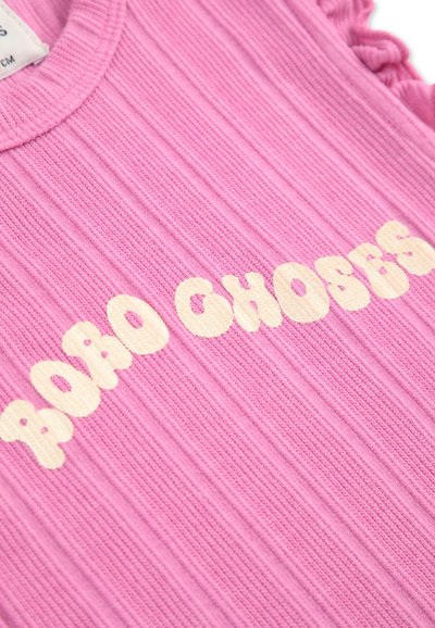 Top de tela acanalada de color rosa con letras redondeadas en blanco que dicen "POD CHOSES". Los volantes en los hombros añaden textura al diseño.