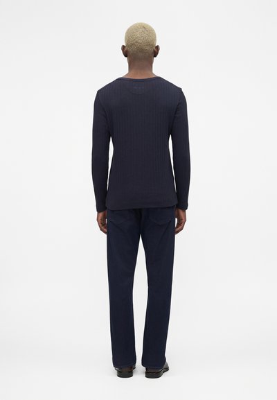 Άτομο με κοντά ξανθά μαλλιά που φοράει μπλε ριμπ μακρυμάνικο τοπ, μπλε παντελόνι και μαύρα παπούτσια, στέκεται γυρισμένο πλάτη σε λευκό φόντο.