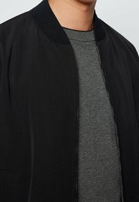 Czarna kurtka typu bomber z gładkiego materiału, z prążkowanym kołnierzem i bocznymi kieszeniami. Noszona na szarym swetrze z okrągłym dekoltem. Proste, czyste linie.