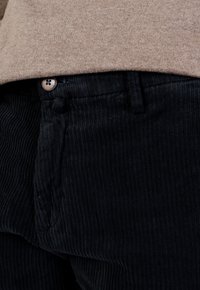 Schwarze Cordhosen mit vertikal geripptem Muster, ausgestattet mit einem Knopf in der Taille und einem flachen Frontdesign. Dezente Nähte sind sichtbar.