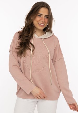 Young woman smiling, wearing a pink hoodie with white drawstrings and white pants, standing against a plain white background.