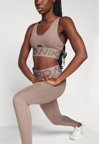 Smeđi sportski crop top i uske tajice s logotipom. Tajice imaju prilagođeni dizajn i glatku teksturu. Bijele tenisice upotpunjuju odjevnu kombinaciju.