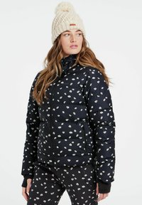 Schwarze Daunenjacke mit kleinem weißem Blumenmuster, Reißverschluss, hohem Kragen und zwei Seitentaschen. Getragen mit passenden Hosen und einer gestrickten cremefarbenen Mütze.
