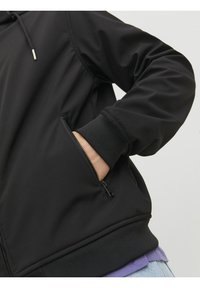 Czarna kurtka o gładkiej teksturze, z zamykaną na zamek kieszenią boczną, dopasowanymi mankietami i jednolitym kolorem, noszona na fioletowej koszuli.