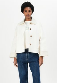White cropped jacket with a pointed collar, featuring large brown buttons and wide sleeves, paired with a light-colored sweatshirt and blue jeans.