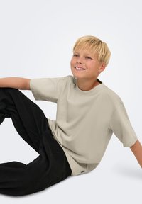 Camiseta de algodón color beige claro con cuello redondo y mangas cortas, combinada con pantalones negros. La tela parece suave y texturizada.