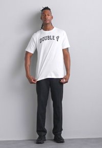 Balta marškinėliai su juodu užrašu "DOUBLE", derinami su juodomis kelnėmis. Modelis stovi tiesiai, rankomis laikydamas marškinėlius, tekstūruotas fonas.