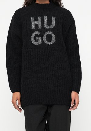 Schwarzer grob gestrickter Pullover mit „HUGO“ in großen weißen, strukturierten Buchstaben auf der Brust, getragen mit schwarzen Hosen.