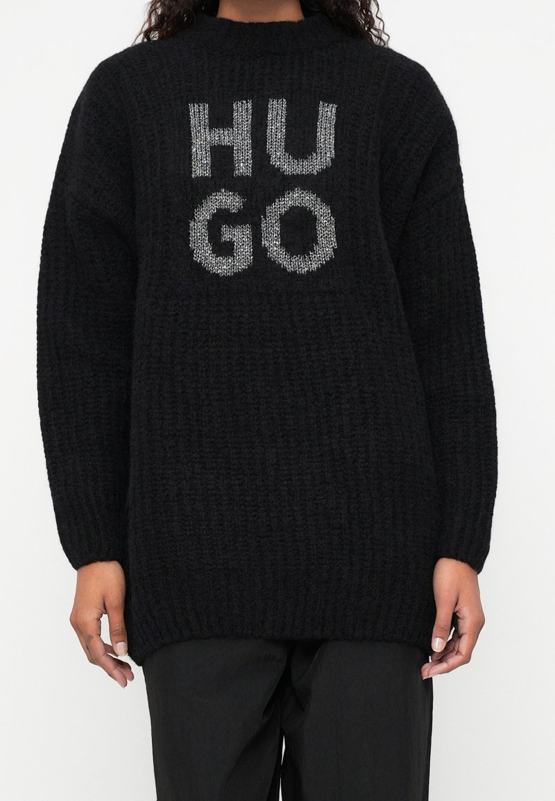 Pull en maille épaisse noire avec "HUGO" en grandes lettres blanches texturées sur la poitrine, porté avec un pantalon noir.