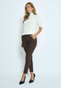 Balts īsām piedurknēm turtlenecks tops, kas apvienots ar brūnām pieguļošām biksēm ar teksturētu audumu un klasisku pogu aizdari. Melni augstpapēžu apavi.
