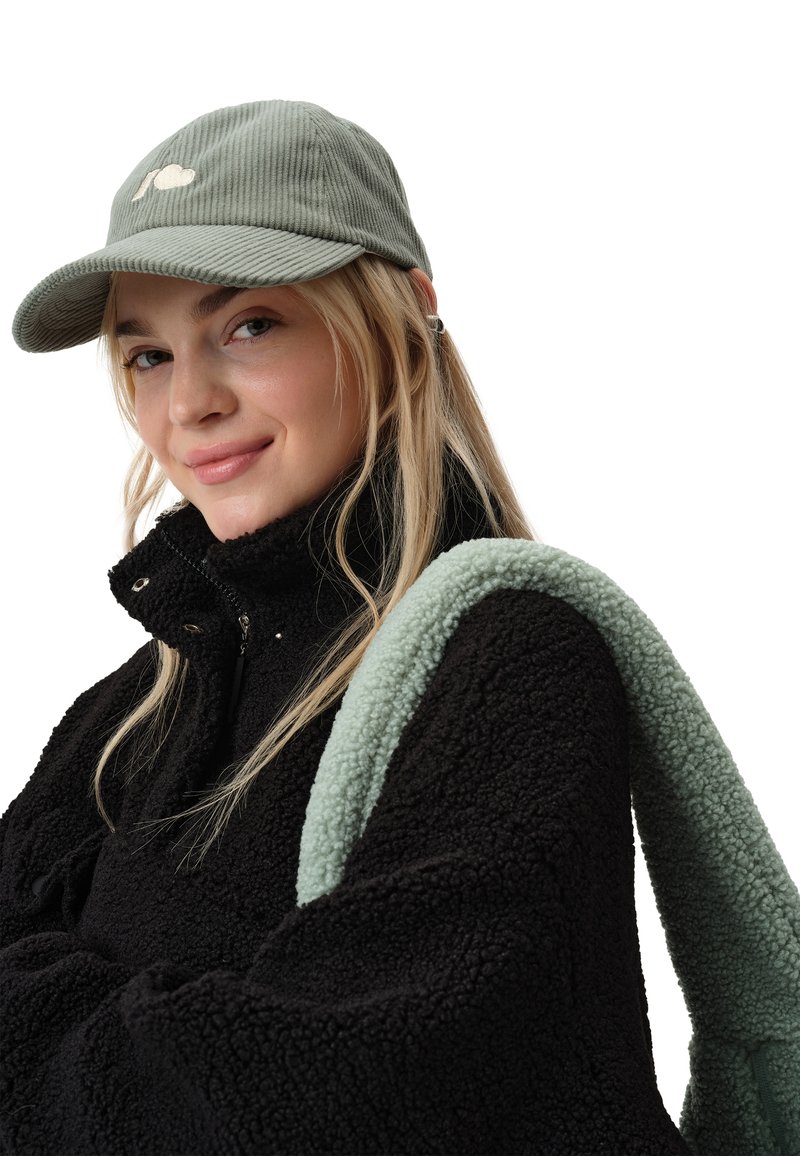 Czarna polarowa kurtka z wysokim kołnierzem, zielona bawełniana czapka z białym logo oraz jasna zielona puszysta ramiączko torby. Modelka ma długie blond włosy.