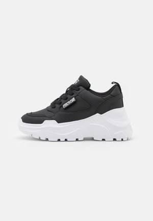 Czarny masywny sneaker z grubą białą podeszwą, czarnymi sznurowadłami i metką Versace Jeans Couture na boku oraz języku.
