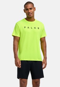 Mężczyzna ubrany w jasny żółty t-shirt z krótkim rękawem z napisem "FALKE" oraz czarne spodenki, stojący na jasnym, jednolitym tle.