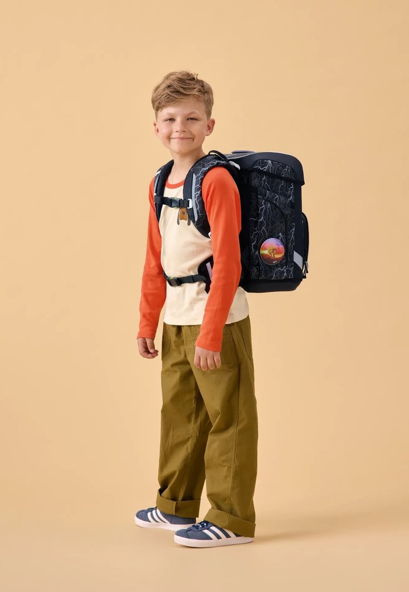 Sac à dos en matière texturée noire, avec des accents orange et jaune, porté par un enfant vêtu d'un t-shirt orange à manches longues et d'un pantalon vert.