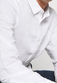 Camicia bianca in lino con colletto classico, caratterizzata da una consistenza morbida e cuciture visibili. Alcuni bottoni sono parzialmente nascosti.