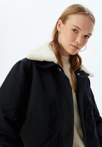 Schwarze Jacke mit einem weichen weißen Lammfellkragen, Knopfverschluss und Seitentaschen. Der Stoff erscheint glatt mit einer strapazierfähigen Textur.