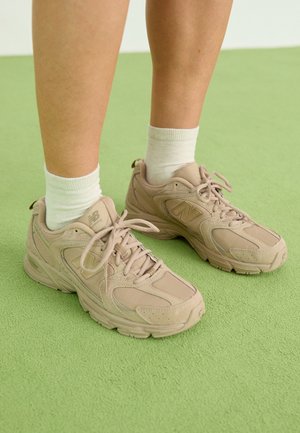 Jalat, joissa on valkoiset sukat ja beiget New Balance -lenkkitossut, seisovat vaaleanvihreällä matolla.