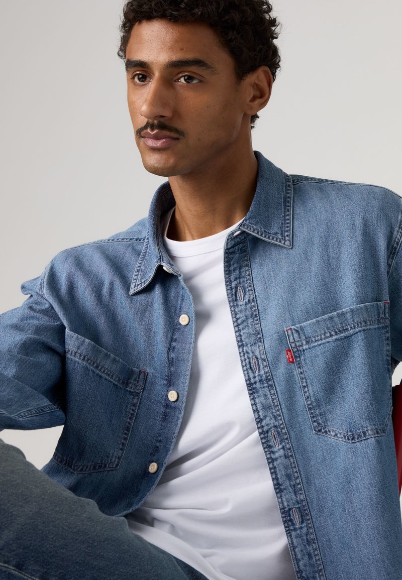 Homme aux cheveux courts et bouclés avec une moustache, portant une chemise en jean bleue par-dessus un t-shirt blanc, assis devant un fond neutre.