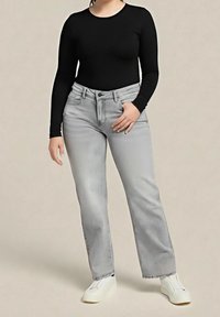 Femme portant un haut noir à manches longues, un jean gris clair à jambes droites et des chaussures blanches sans lacets, debout devant un fond beige uni.