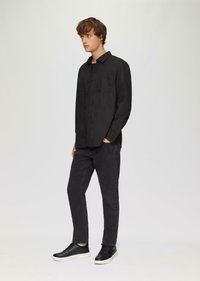 Zwart overhemdd met een enkele borstzak, gecombineerd met donkergrijze slim-fit jeans en zwarte high-top sneakers, tegen een lichte achtergrond.
