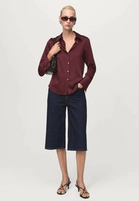 Woman wearing dark red button-up shirt, dark denim culottes, black heeled sandals, sunglasses, and holding a black handbag over one shoulder.