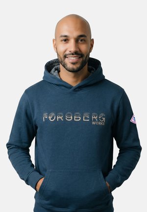 Uomo calvo sorridente con barba che indossa una felpa con cappuccio blu navy Forsberg Works con le mani nella tasca frontale su uno sfondo chiaro uniforme.