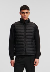 Vesta puffer neagră cu guler înalt și închidere cu fermoar, având un design matlasat și buzunare laterale. Purtați peste o cămașă neagră cu mânecă lungă.