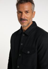 Jachetă din lână neagră cu guler, butoane frontale și un buzunar pe piept cu închidere prin buton. Textură moale și croială ajustată.