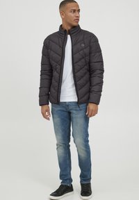 Jachetă puf cu model în zigzag, guler înalt și închidere cu fermoar. Pur tată pe un tricou alb și asortată cu blugi albaștri și adidași negri.