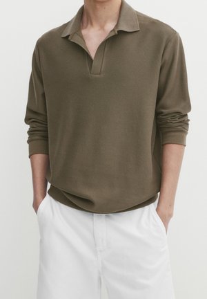 Olivgrünes Poloshirt mit langen Ärmeln, V-Ausschnitt, geripptem Muster und klassischem Kragen. Kombiniert mit weißen Hosen.