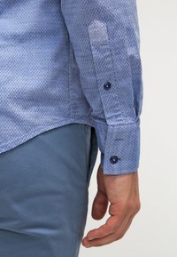 Modrá vzorovaná košile s dlouhým rukávem a malými diamantovými tvary, s manžetou na knoflík a hladkou texturou, doplněná o světle modré kalhoty.