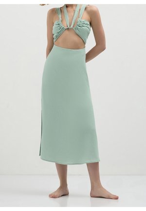 Vrouw die een lichtgroene jurk draagt met een uitgesneden taille en geplooide bandjes, staat blootsvoets tegen een effen witte achtergrond.