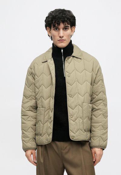 Μπεζ γούνινο μπουφάν με διαμαντένιο σχέδιο, μπροστινές τσέπες, κουμπιά με σνάπ και γιακά. Φορεμένο πάνω από έναν μαύρο πουλόβερ με ψηλό λαιμό.