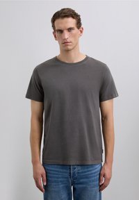 Un homme avec des cheveux courts et bouclés portant un T-shirt à manches courtes gris foncé uni et un jean bleu, se tenant contre un fond gris clair.