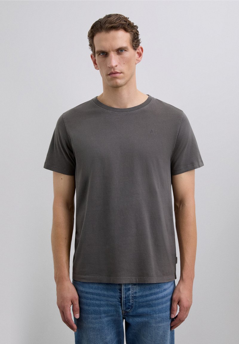 Un homme avec des cheveux courts et bouclés portant un T-shirt à manches courtes gris foncé uni et un jean bleu, se tenant contre un fond gris clair.