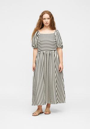 Jeune femme aux longs cheveux roux portant une robe longue rayée noire et blanche avec des manches bouffantes et des sandales beiges, debout sur fond blanc.