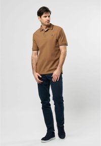 Hnědé polo tričko s námořnickým kontrastním límcem, v kombinaci s tmavě modrými džíny. Model stojí uvolněně, s rukama na bocích.