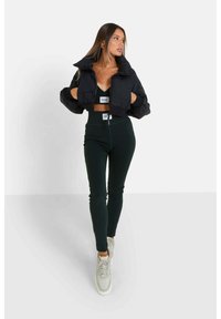 Mujer con una chaqueta de plumas negra corta, top negro corto, leggings de cintura alta con canalé y zapatillas blancas, de pie contra un fondo blanco.