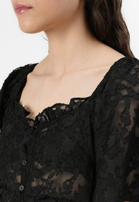 Black lace blouse featuring a square neckline, button closure, and textured floral pattern. Lightweight and semi-sheer fabric.