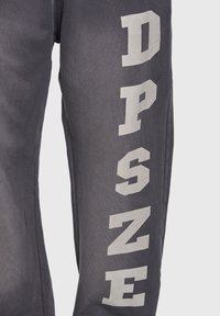 Pantalon de survêtement gris foncé avec de grandes lettres blanches audacieuses formant le mot "DPSNE" verticalement le long du côté extérieur d'une jambe.