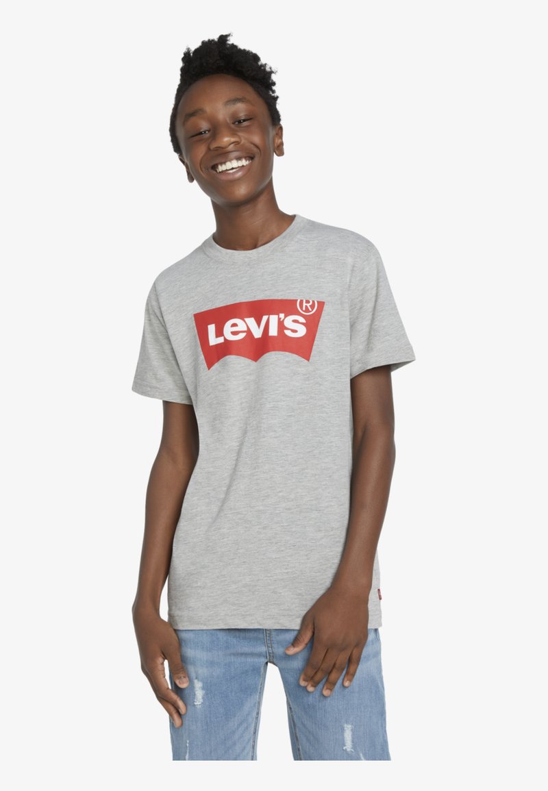 Camiseta de algodón gris con un destacado logotipo rojo de Levi's. Mangas cortas, cuello redondo y un corte relajado. Combinada con jeans azules desgastados.