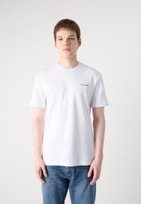 Fehér, rövid ujjú pamut póló, kerek nyakkal, amelyen egy kis fekete "Calvin Klein" logó található a mellrészen, kék farmernadrággal párosítva.