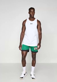 Camisola branca sem mangas com logo preto, calções verdes com um logo e ténis brancos com detalhes pretos. Modelo a posed sobre um fundo neutro.