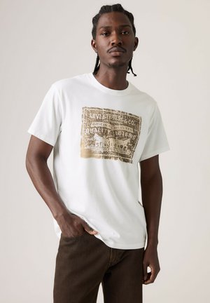Man met gevlochten haar, gekleed in een wit T-shirt met een vervaagde bruine afbeelding en een bruine broek, staand met één hand in de zak.