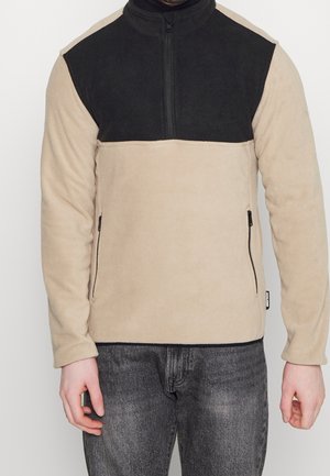 Mężczyzna ubrany w beżowo-czarny polar z kieszeniami na zamek, w połączeniu z ciemnoszarymi dżinsami, na jasnym, jednolitym tle.